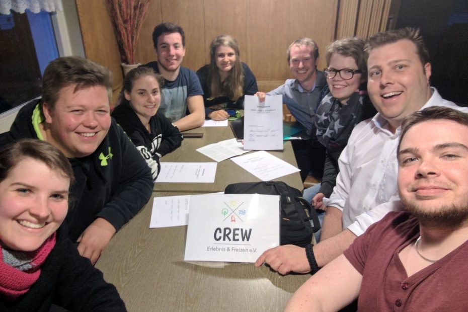 Eine Gruppe junger Menschen sitzt an einem Tisch; einer hält das CREW-Logo hoch, ein anderer das Gründungsprotokoll des Vereins