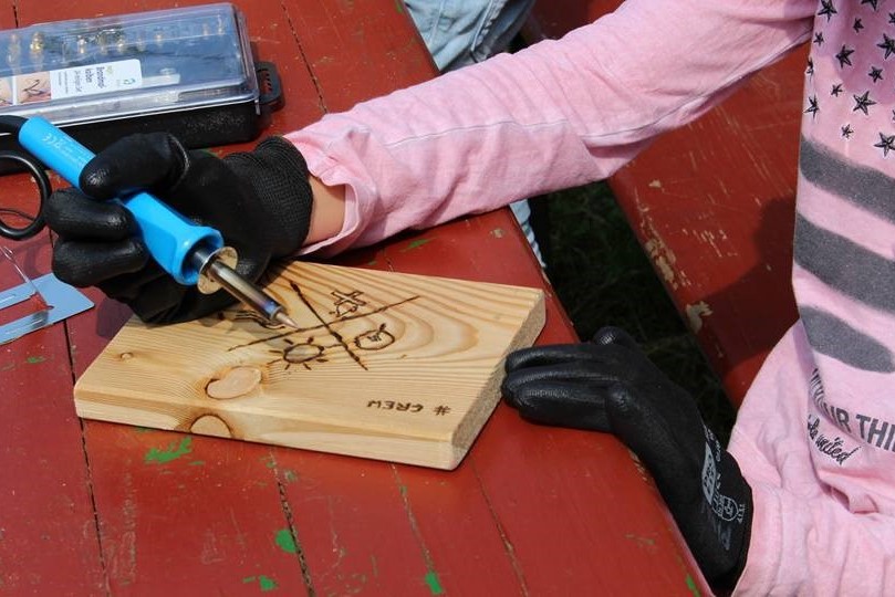 Eine Hand führt einen Lötkolben und brennt das CREW-Logo in ein Stück Holz