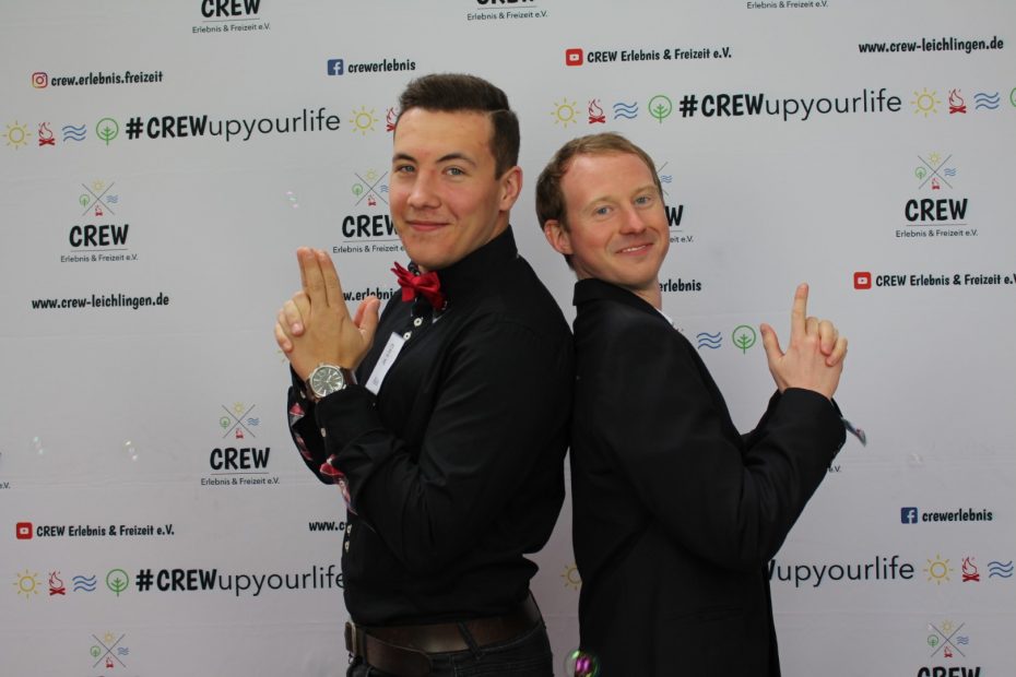 Zwei CREW-Ehrenamtler posieren im Anzug vor einer CREW-Messewand