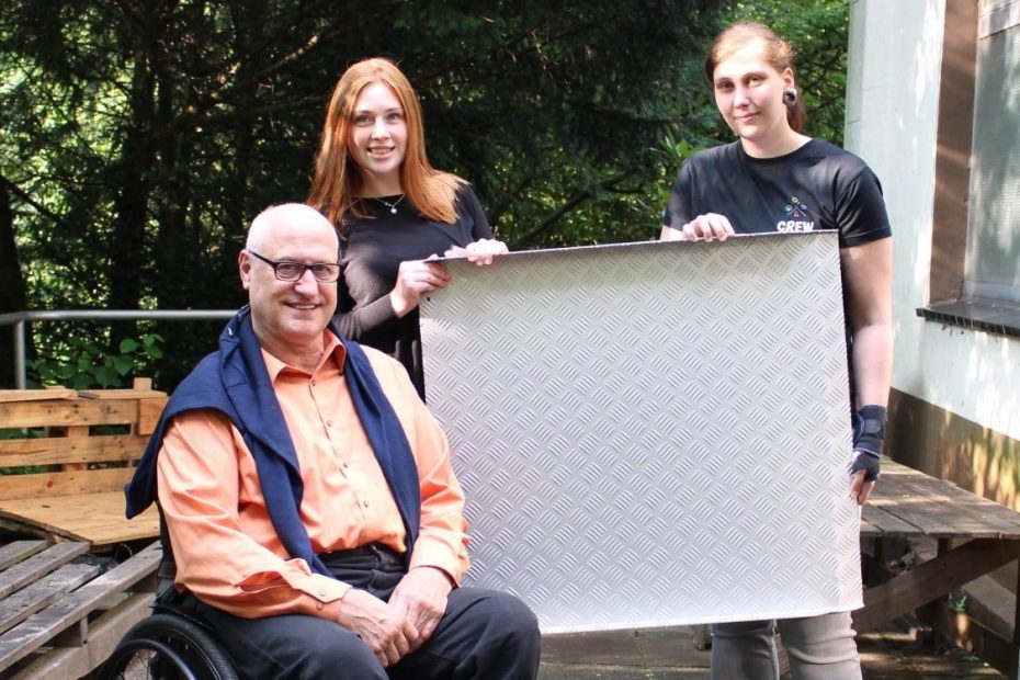 Zwei CREW-Ehrenamtlerinnen bekommen von einem Herrn im Rollstuhl eine Rollstuhlrampe als Spende überreicht